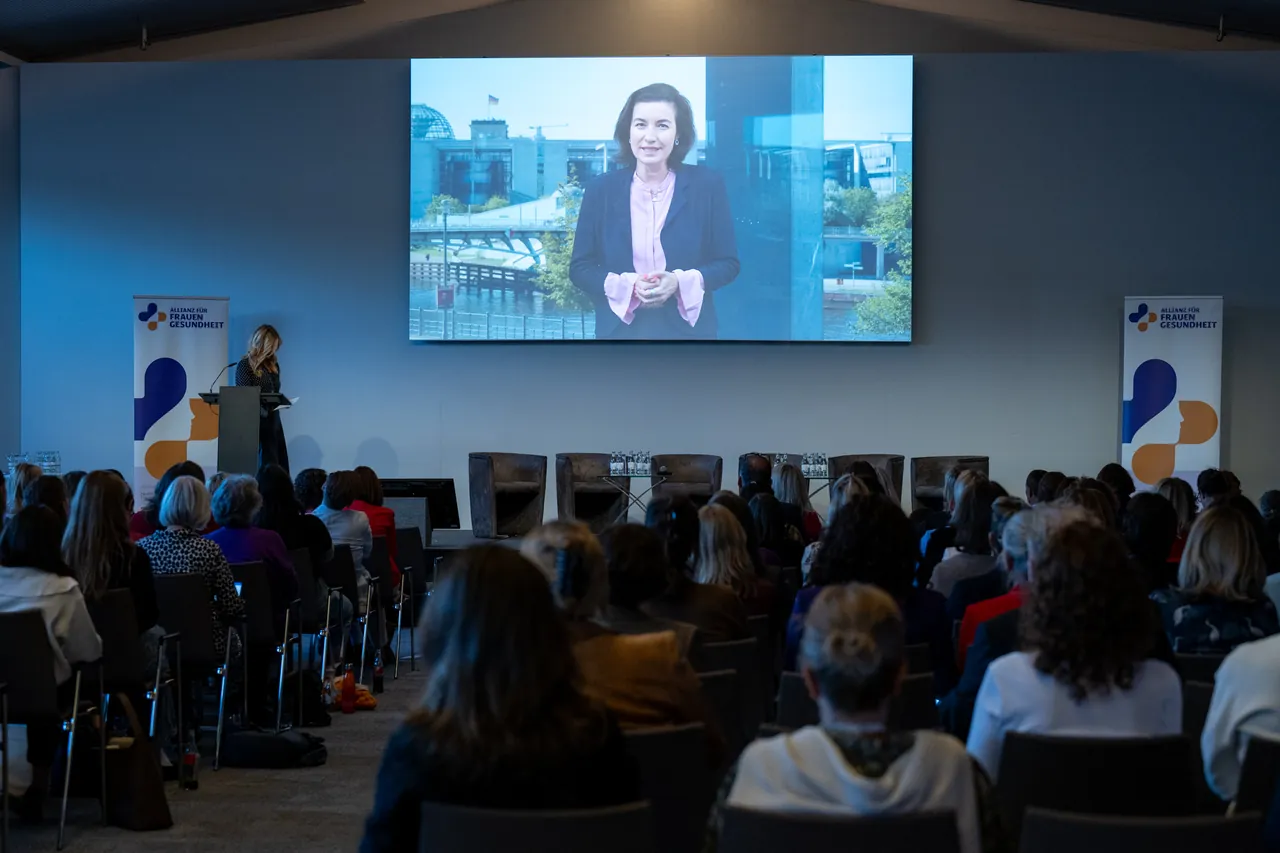 Videobotschaft einer Frau auf der Leinwand vor vollem Saal